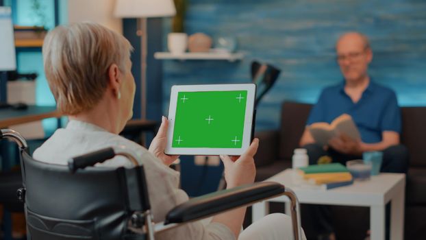 Adult in wheelchair holding digital tablet with green screen. Senior woman with chronic disability looking at isolated copy space with blank mock up background and chroma key template.
