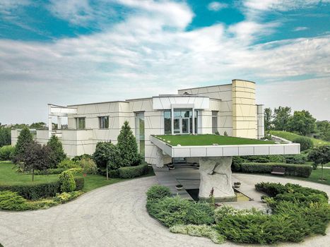 Luxurious modern country house with roofed porch at main entrance and massive decorative stone column supporting roof, surrounded by green parkland in cloudy summer day