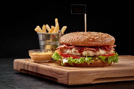 Tasty big burger with chicken patty, fried bacon slices, grated parmesan and fresh vegetables in fluffy sesame bun with french fries and aioli sauce on wooden serving board in fast food restaurant