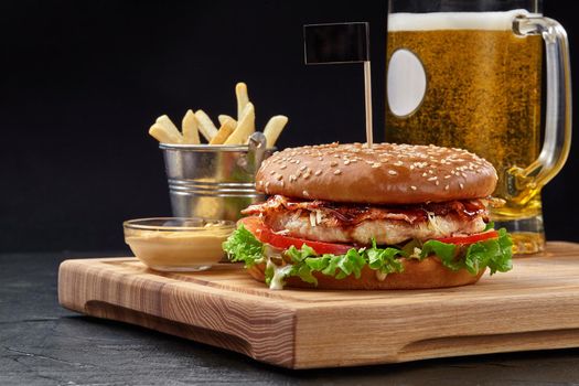 Tasty burger with chicken patty, fried bacon, grated parmesan and vegetables in sesame bun served with glass of foamy beer, crispy fries and aioli sauce on wooden board. Popular fast food dish