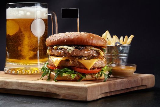 Hamburger with two juicy patties, emmental slices and blue cheese, caramelized onions, fresh tomatoes and arugula in sesame bun served with glass of pale lager, french fries and sauce on wooden board