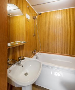 Interior of small bathroom in hotel room with walls faced with wooden slats, equipped with white wash basin and acrylic bathtub with chrome fixture and mirror