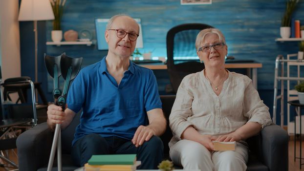 POV of retired couple waving at videoconference webcam in living room. Senior man and woman with physical disability talking to people on remote teleconference call, using online connection.