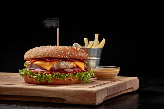 Delicious cheeseburger with pork and veal patty, fried bacon, fresh red onion slices, tomatoes and lettuce in fluffy bun sprinkled with sesame served with crispy fries and aioli sauce on wooden board