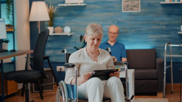 Aged woman using digital tablet while she sits in wheelchair, holding gadget with touch screen to browse internet. Grandmother dealing with physical disability and looking at device.