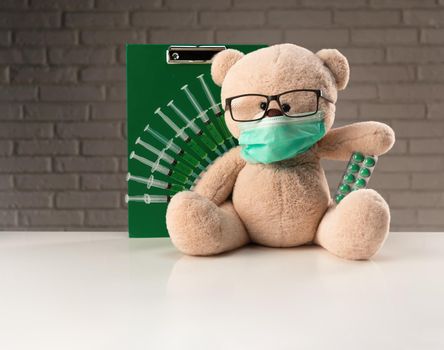 teddy bear in a medical mask with syringes in his shoulder on a white medical table with pills