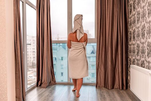 Back view of a young woman wrapped in a towel standing in front of the wondow. Young woman after morning shower