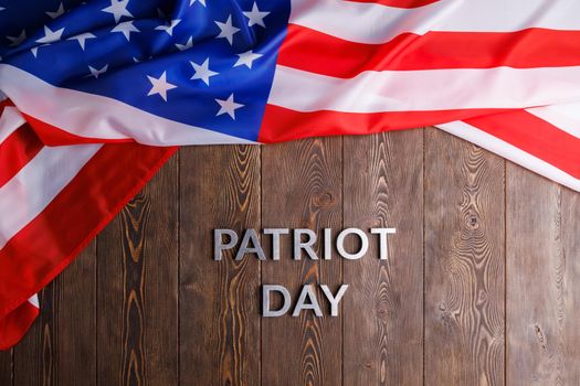 the words patriot day laid with silver metal letters on wooden board surface with crumpled usa flag above, in flat lay high angle directly above view