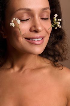 Serene delighted pretty natural beauty young woman with closed eyes and beautiful toothy smile posing with Gypsophila sprigs on her face. Skin care and body positivity concept. Cosmetology dermatology