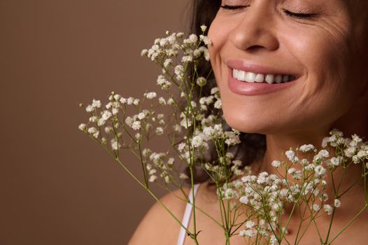 Close-up portrait of Hispanic pretty woman with closed eyes and beautiful smile, holding Gypsophila white sprig against beige background. Femininity, sensuality, natural beauty, Women's Day concept