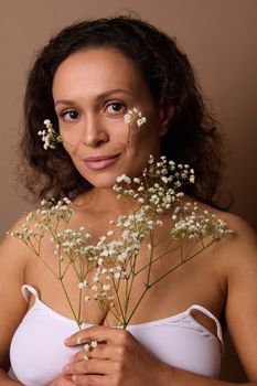 Fashion beauty portrait of attractive sensual feminine woman with Gypsophila white sprig confidently looking at camera, posing against beige colored background. Body, skin care, Women's Day concept