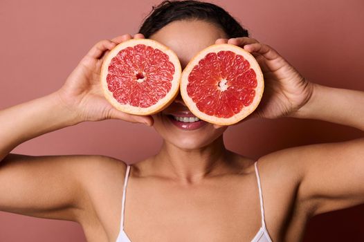 Attractive young woman covering her eyes with two halves of fresh juicy grapefruit, smiling with beautiful toothy smile, posing against pastel coral background. Skin and body care concept. Copy space