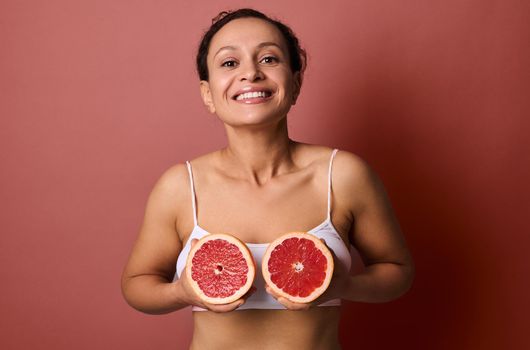 Attractive woman in a white underwear holds grapefruits halves at chest level, smiling sweetly with a toothy smile posing on a coral background with space for text and advertising. Body and skin care