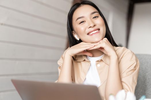 Cute smiling asian girl working from home on laptop, watching video with earphones on computer, doing homework or video chatting.