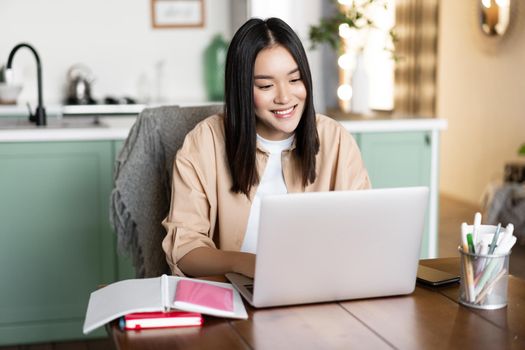 Woman having video chat, online school on computer, smiling at laptop camera. Girl studying remotely from home, sitting in kitchen with books and computer.