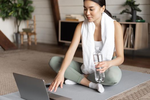 Young asian woman connect to online fitness instructor on laptop, workout via video chat at home, sitting on floor mat.