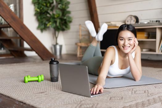 Asian fitness girl taking online fitness course, watching sports video, exercising at home on floor mat with dumbells and protein shaker bottle, smiling at camera.