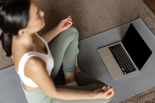 Cropped shot of asian woman meditating in floor mat at home, listening to meditation podcast on laptop, practice yoga online courase, wearing activewear.