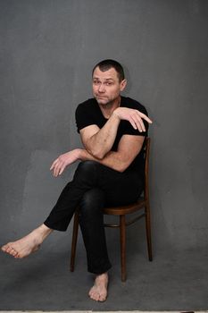 Full length photo of a man sitting on a chair with his hands folded and looking at the camera with emotion. Vertical photo on a gray background