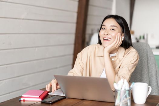 Happy asian girl listening online webinar, attend internet course classes on laptop, wearing wireless earphones and smiling, working from home.