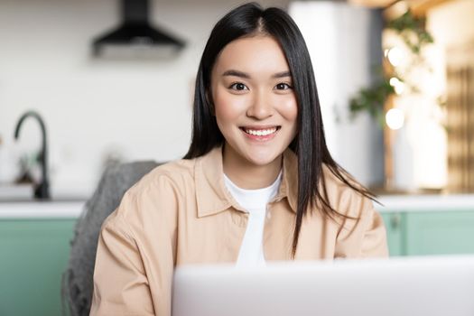 Freelance asian girl sitting at home and working with laptop, smiling at camera. Young woman using computer, studying remote, taking online course.