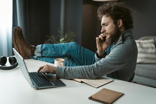 Brutal Businessman has a Serious Telephone Conversation with Suppliers and is Typing on a Computer. Feet on the Table. Work Desk with Laptop, Headphones, Diary and White Cup. Male Profile. Copy Space. Close-up. High quality photo