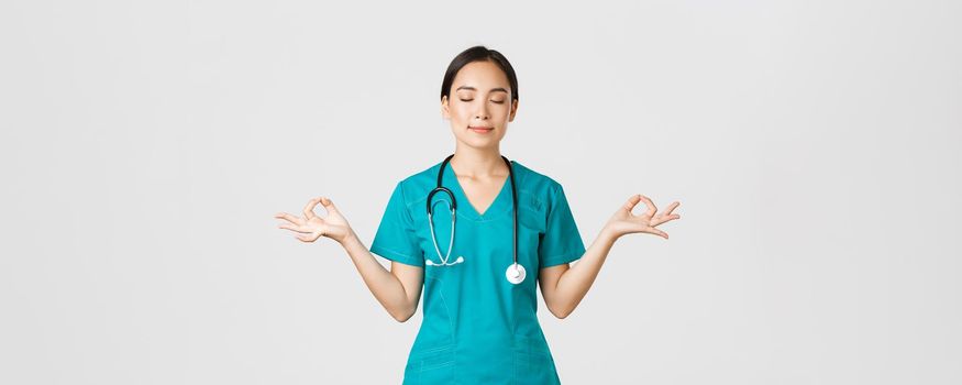 Covid-19, healthcare workers and preventing virus concept. Calm and patient asian female doctor, nurse in scrubs staying relaxed, meditating with eyes closed and happy smile, white background.