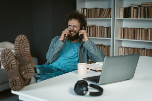 Male Model Poses with Feet on the Table and Smartphone in his Hand.There are Laptop, Mug of Tea, Diary and Earphones on the White Desk . Shelves Background. Close-up . High quality photo