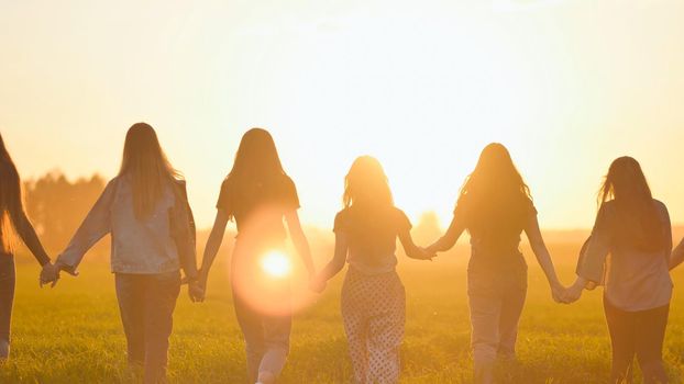 A group of girls walk towards the sun at sunset holding hands