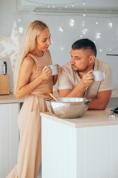 Caucasian couple drinking hot chocolate while preparation dough for Christmas cookies in the decorated kitchen at home. Christmas party preparation. Couple talking while drinking hot cocoa.