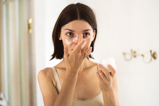A young girl applies a moisturizing cream mask near the mirror in the bathroom and holds a jar of balm in her hand. A woman takes care of the health of her skin, uses cosmetic masks.