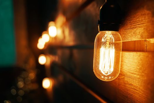 Holiday interior decorating. Antique incandescent Edison type bulb on wooden texture wall. Blurred warm garland light on background. Christmas night party mood. Copy space for congratulations.