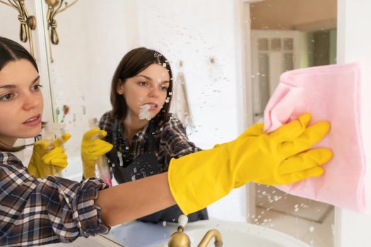 A young girl is cleaning the bathroom, applying detergent with a spray and washing the mirror with a sponge in yellow gloves on her hands. Smiling woman taking care of the cleanliness of her home.