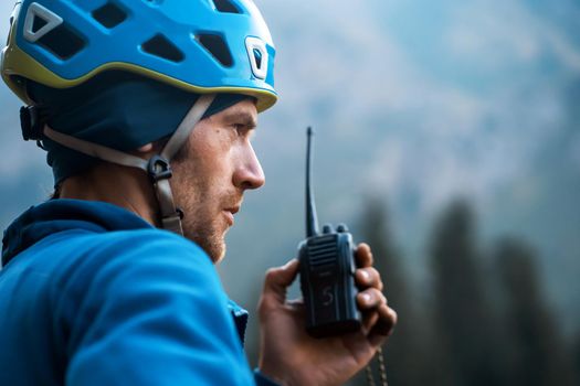 A man in a helmet and a windbreaker is talking on a walkie-talkie against the background of the mountains, a mountain rescuer checks the connection with the base, close-up view.