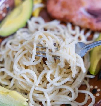 Extreme close-up of fork in white creamy spaghetti surrounded by avocado slices. Pasta with cream and pork dish on traditional brown plate. Cuisine and balanced diet