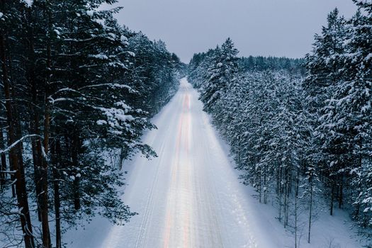 Night time aerial perspective view of snowy road in pine tree forest in winter season. Drone view of snowy winter road surrounded pine and fir forest and light trace of car