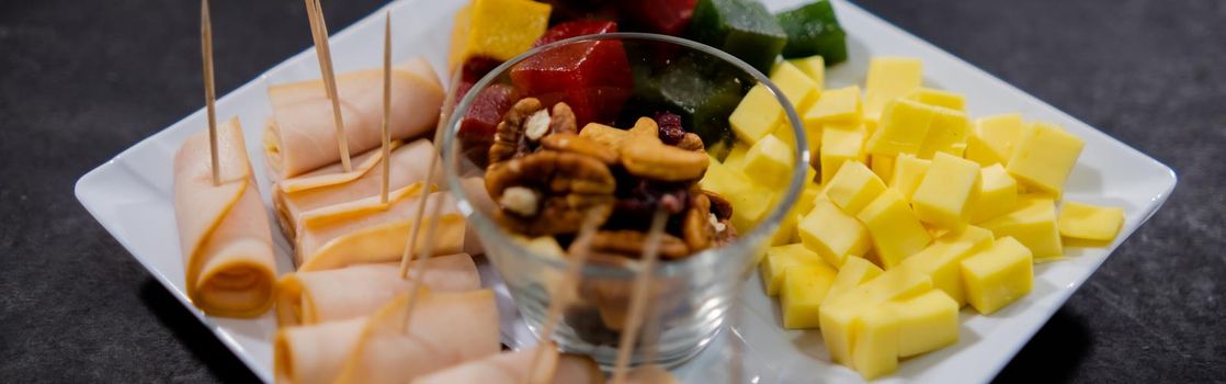 Wide view of turkey ham rolls, diced fruit paste, cheddar cheese cubes, and glass of walnuts on square white plate. Turkey meat, colorful Mexican candy, and nuts above black surface. Healthy snacks