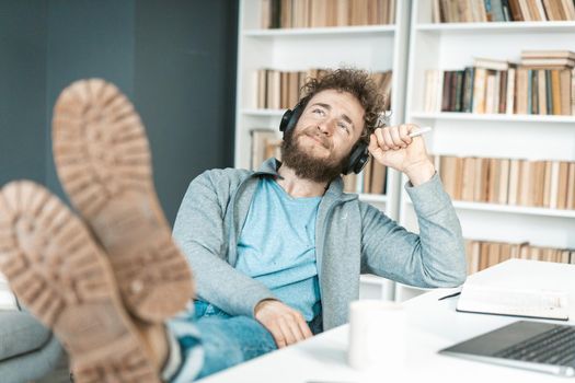 Casual Guy Listens to Relaxing Music on Headphones in his Office, Feet on the Table, Soles to the Camera. Bookshelves Background. Close-up. High quality photo
