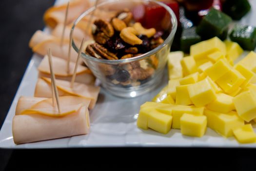Close-up of turkey ham rolls, diced fruit paste, cheddar cheese cubes, and glass of walnuts on square white plate. Turkey meat, colorful Mexican candy, and nuts above black surface. Healthy snacks