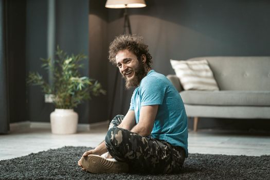 Young Guy Sits on the Carpet with Folded Legs. Curly-haired Man in a Blue T-shirt Siting on the Floor and Laughing .Copy Space, Close-up. High quality photo