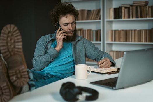 Caucasian Man has a Telephone Conversation and Takes Notes in his Diary. Feet on the Table. Soles to the Camera. Shelves Background. Close-up. High quality photo