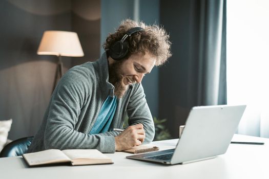 Young Student has an Online Video Lecture Uses a Laptop and Headphones, Takes Notes in a Notebook. Curly-Haired Guy sits in Earphones at the Table in his Office. Close-up. High quality photo