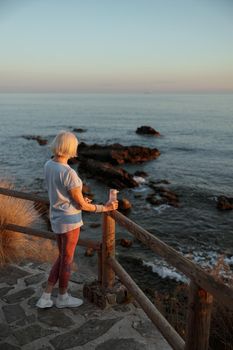 Back view of athletic elderly female in sportswear, standing at wooden fence and enjoying beauty of wild nature