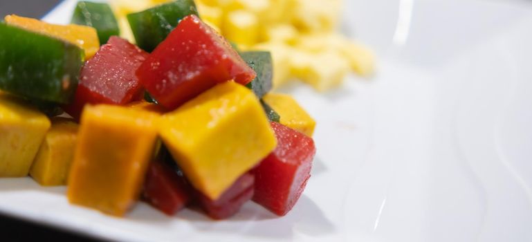 Extreme close-up of red, yellow, and green fruit paste cubes and diced cheese on white plate. Porcelain plate of fresh yellow cheese slices and traditional fruit candy. Sweet and healthy snacks