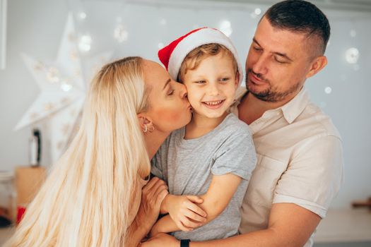 Loving parents hugging their little son in Santa hat at home with decorated garland or lighting lantern on Christmas Eve. Family celebrating Christmas and New Year together.