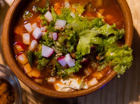 Top view of clay bowl of delicious and traditional red pozole on wooden table. Authentic Hispanic pork stew in handmade bowl with chopped radish and lettuce on top. Traditional Mexican cuisine