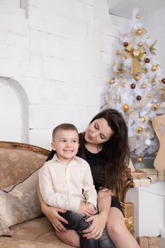 Atmosphere of coming holidays. portrait of happy single mother with her little pleasant boy in white bright room near christmas tree in golden tones.