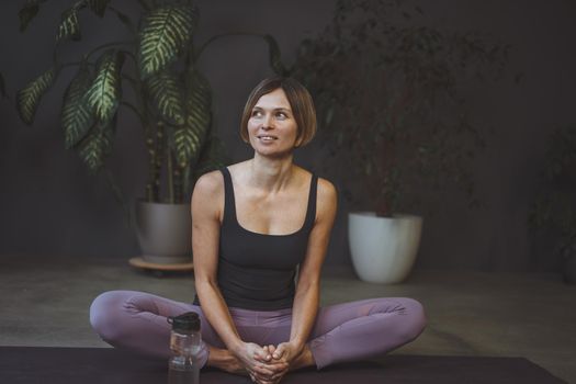 Happy young woman after a home yoga workout sits on a mat with a water bottle. Butterfly Pose. Concept of a healthy lifestyle, sports, care, longevity. High quality photo