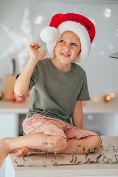 Happy little boy in a Santa hat is sitting on the kitchen table and playing with different shaped cookie cutters. Boy ready to cook Christmas cookies for his family. Kids being kids and having fun.