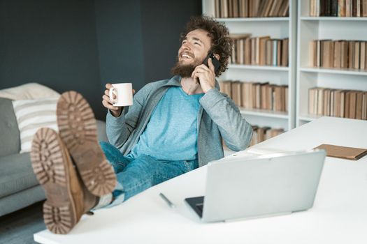 Positive Businessman in Casual Clothes has a Easy Phone Conversation with a Cup of Coffee in his Hand and Feet on the Table. Soles to the Camera. Cute Caucasian Guy Talking on the Smartphone in the Office. Close-up. High quality photo